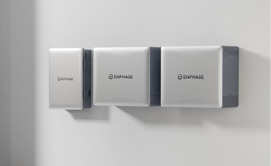 Three Enphase battery systems mounted on a wall with a garden visible outside.
